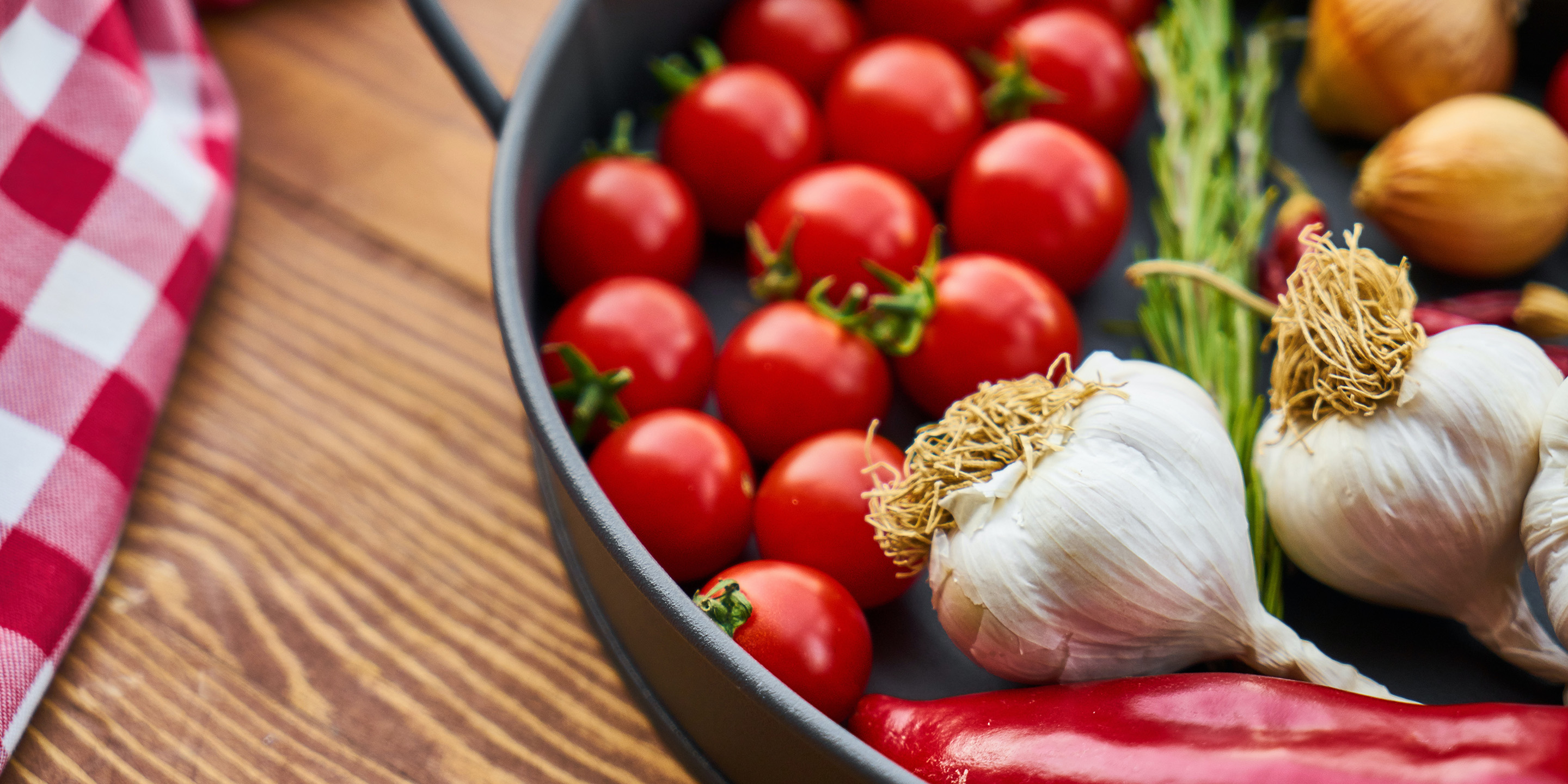 Skillet with bright red grape tomatoes, unpeeled onions and garlic, and red pepper on wooden board with red and white checkered fabric.