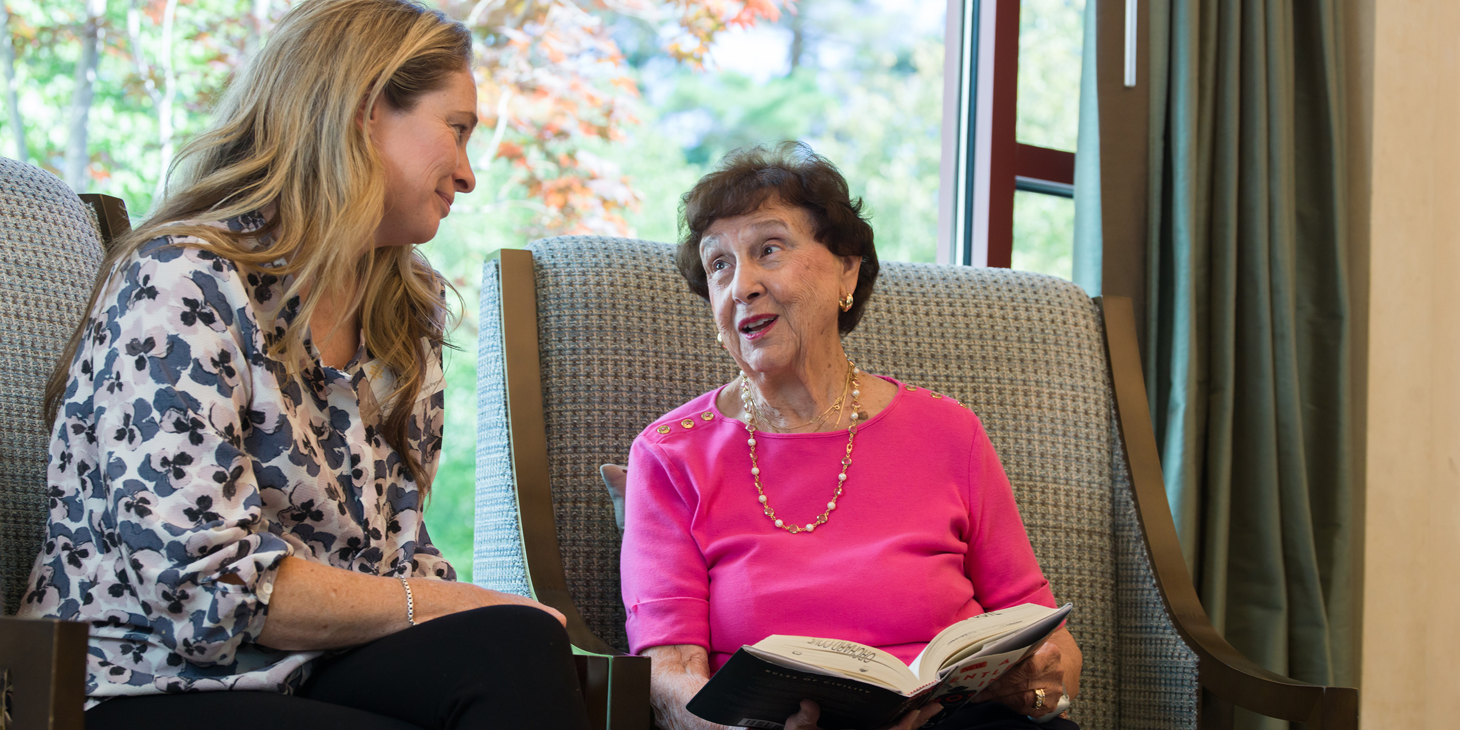 Older woman in pink shirt with a book open on her lap, speaking with a woman.