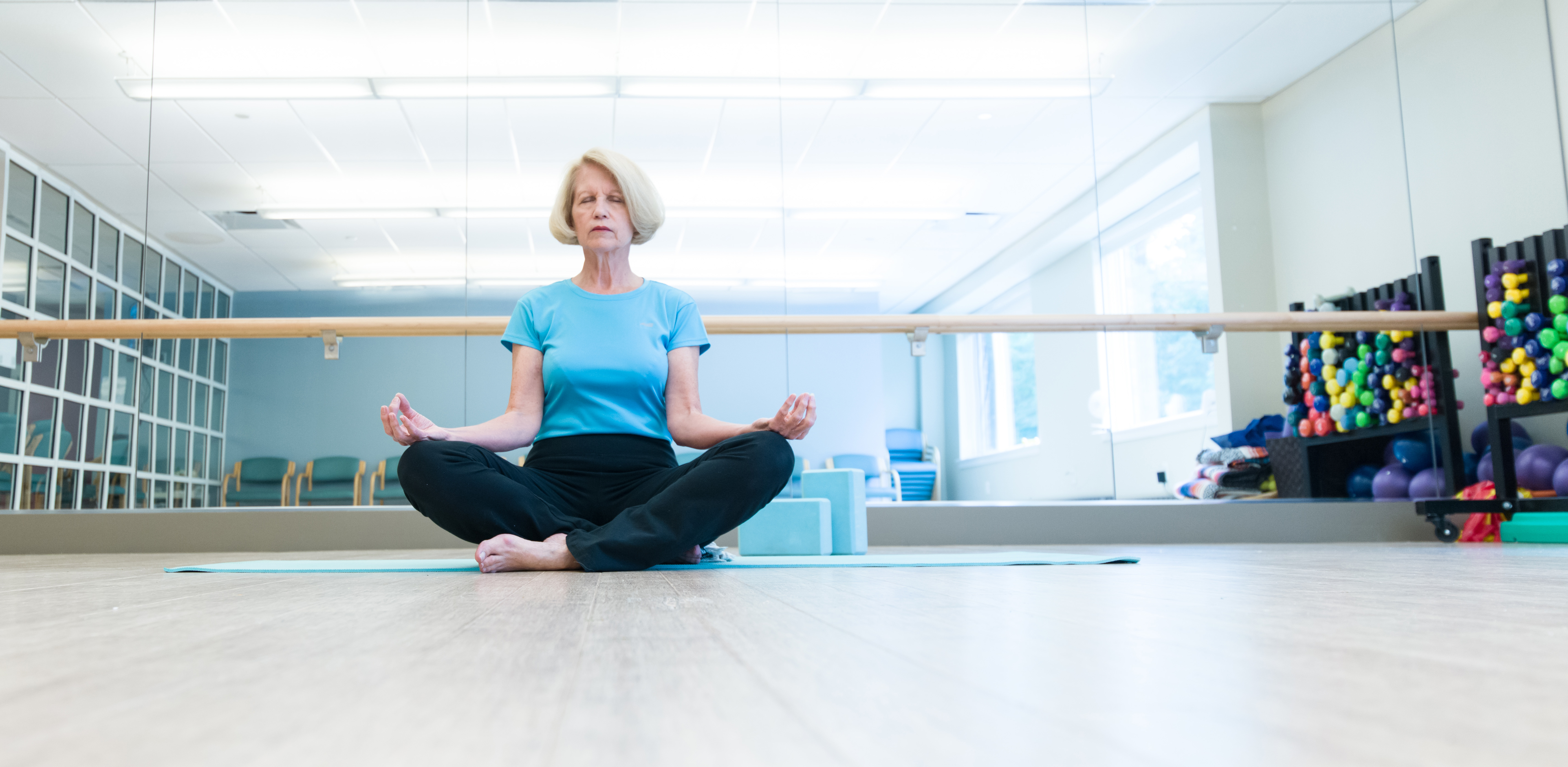 Woman in aqua blue shirt sitting cross-legged, in a meditative pose with the back of her hands on her knees.