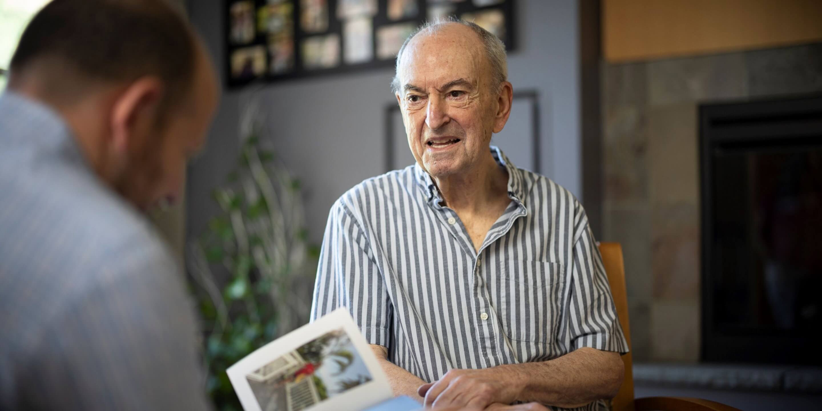Older man sitting while another man is looing at a photo book