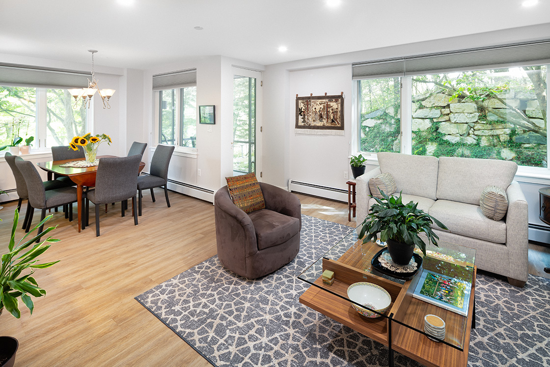 Spacious living room with couch, sitting chair, door out to a porch, and large windows looking out to trees.