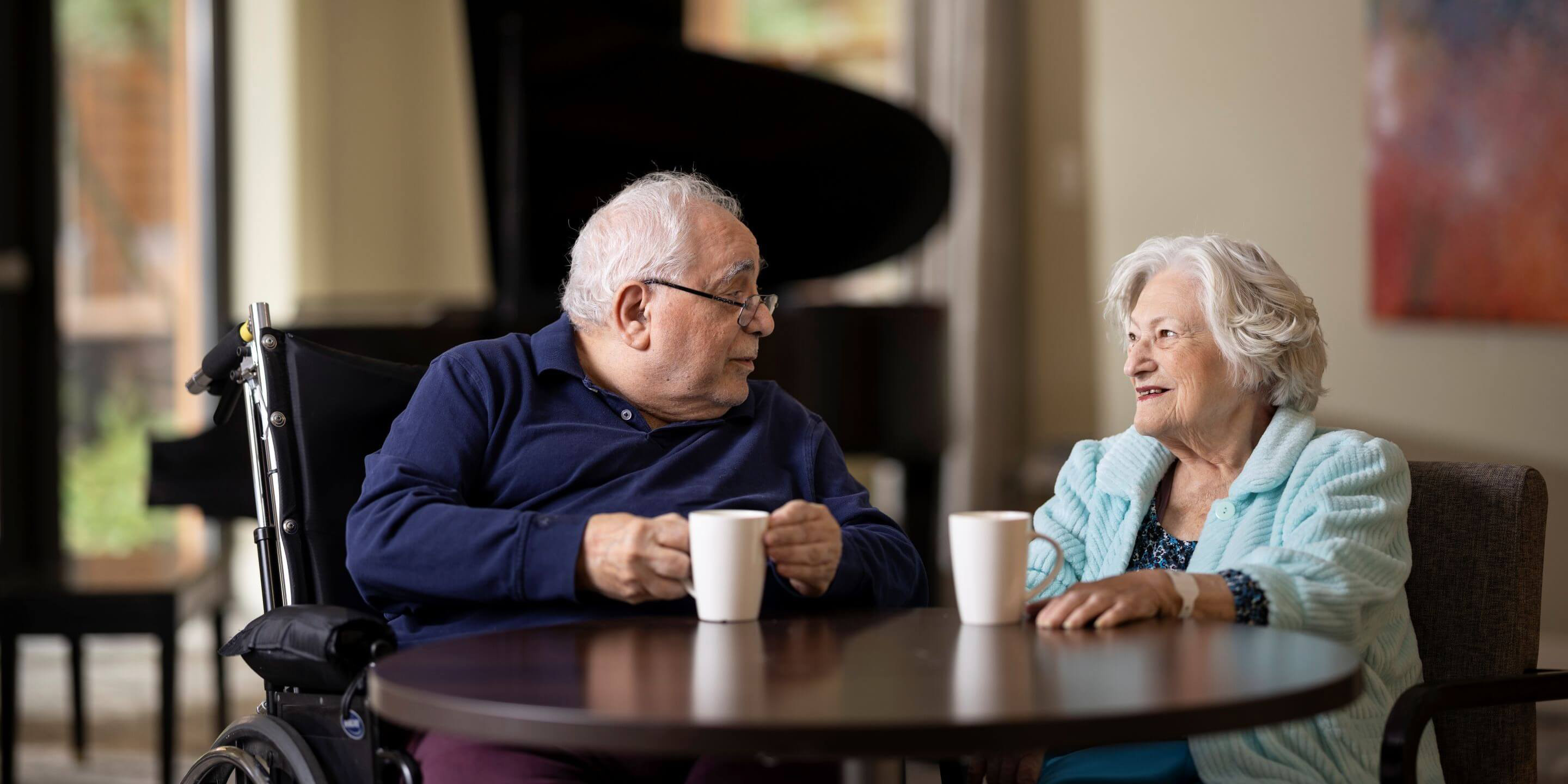 Older man sitting in a wheelchair at a table having coffee with an older woman