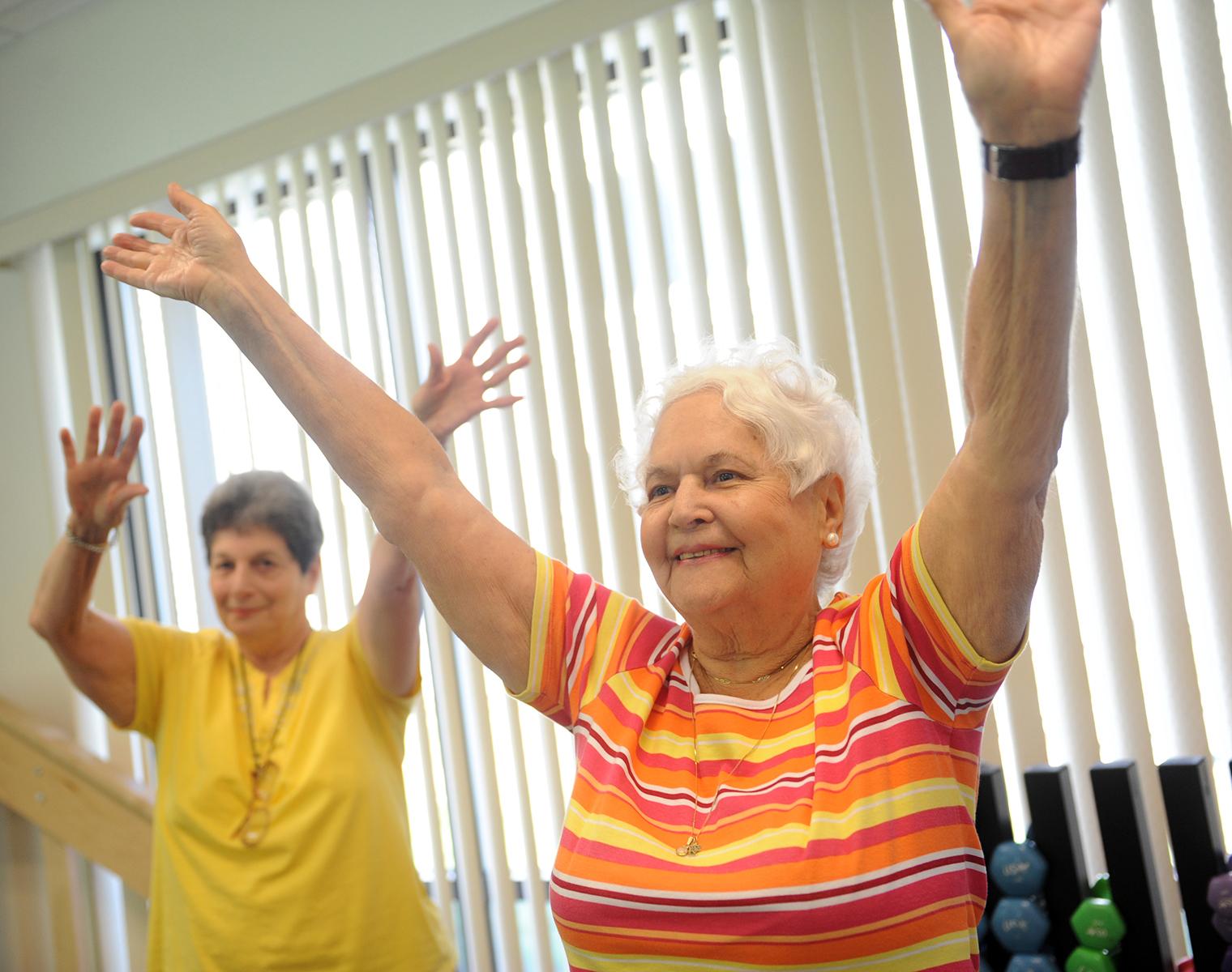 Two smiling older women, one in the background and one in the foreground, hold their arms in the air. Behind them is a wall of windows and a weight rack with dumbells.