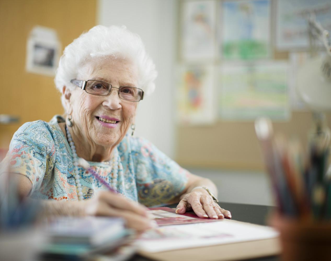 An older woman sits at a table, holding a pencil, sketching on a notepad. There is a jar of colored pencils in the foreground and artwork hanging on the wall behind her.