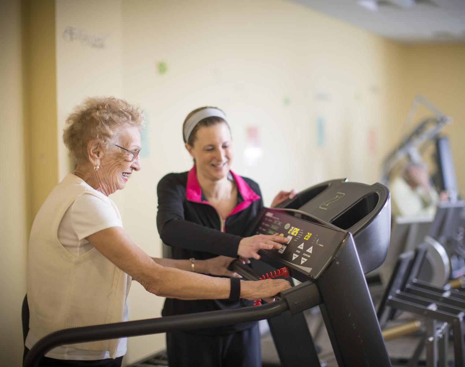 An older woman is walking on a treadmill. A younger woman in workout clothes stands next to her, changing the display on the treadmill. They are both smiling.