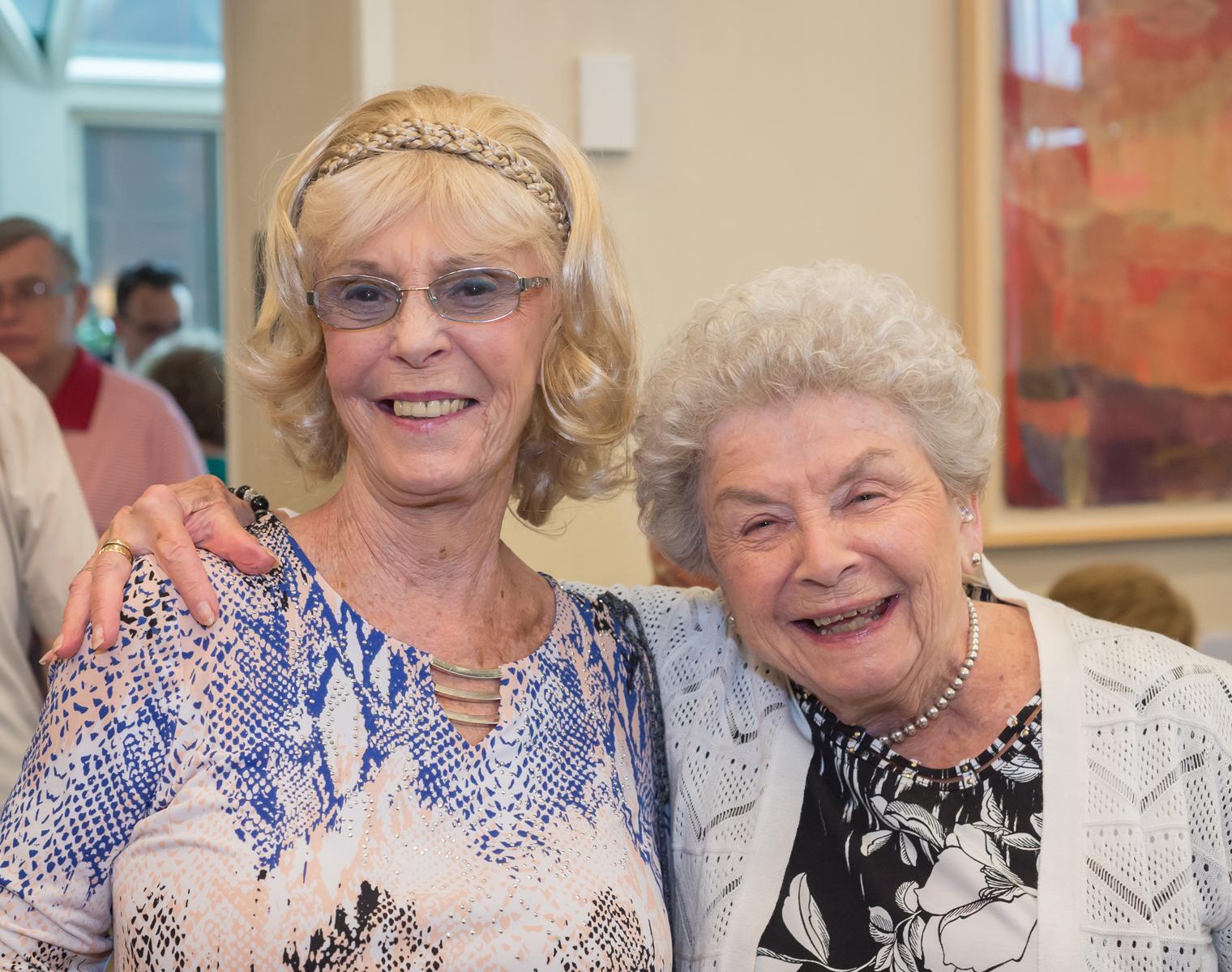 Two older women with big smiles stand close to each other looking at the camera. One woman has her arm around the other.