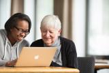 A female volunteer sits with an elder female and helps her use a laptop.