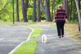 An older adult woman walks on a trail with her dog's leash in one hand and a walking stick in the other.