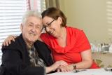 A male senior living resident and his adult daughter embrace in a hug at a dining table.
