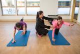 Two women do yoga poses while one gets help from an instructor 