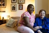 A female caregiver sits on a bed while a female patient sits on a chair next to the bed