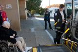 Two employees who work in the Security department at Hebrew SeniorLife help a resident in a wheelchair get loaded onto a lift to get into a transport vehicle 