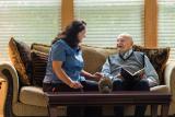 An older male sits on a couch with a woman caregiver and reads.