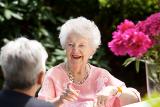 An elder woman wearing a pink shirt sits around a table outside and talks with another woman.