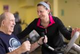 A female trainer helps an elder male while he works out on a stationary workout bike