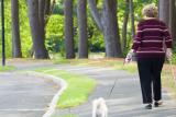 A woman walks down a path holding a dog leash in one hand and a walking stick in the other.