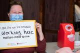 A Hebrew SeniorLife employee sits in a chair, wearing a surgical mask with a sign that says “I got the vaccine because working here matters most to me.” The words “working here” are handwritten.