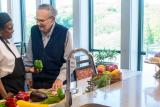 A male patient stands in the kitchen of his household at Hebrew Rehabilitation Center in Boston. A female member of the culinary team stands next to him, holding a green pepper. In front of them there are a variety of colorful fruits and vegetables on the counter. They are looking at each other and smiling.