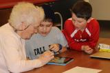An older person and two middle school students sit at a table, looking at a tablet together.