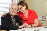 A male senior living resident and his adult daughter embrace in a hug at a dining table.