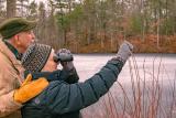 Outdoor winter scene in woods and water with man who has an arm around a woman who is looking through binoculars and pointing.
