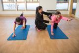 Two women crouching on yoga mats with outstretched arm and leg receiving guidance from a trainer.tw