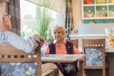 Sitting in a dining area, an older woman in a pink shirt and purple vest talks with a man in a black and white plaid shirt whose back is to the camera.