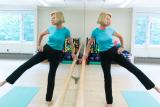 Woman wearing a blue shirt and athletic clothes standing up and holding onto a barre extending her leg to the side for a stretching exercise.