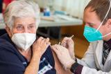 Woman in a blue shirt wearing a mask receives COVID-19 vaccine administered by man with a tie and labcoat.