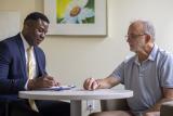 Older man sitting at table with doctor in blue blazer discussing report on clipboard. 