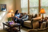 Woman with a hand on the shoulder and holding the hand of an older man sitting on a couch in a living room