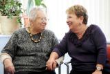 Two women sitting in chairs holding hands, smiling and talking to each other.