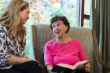 Older woman in pink shirt with a book open on her lap, speaking with a woman.