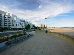 On the left, a white apartment building about eight stories high sits in front of Revere Beach Boulevard. On the right, we see the ocean and a beach, with benches and a sidewalk running alongside.