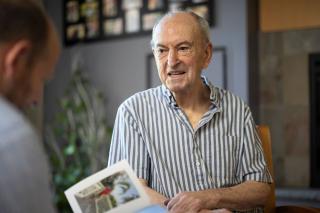 Older man sitting while another man is looing at a photo book