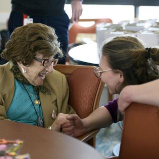 An older woman sits at a table and smiles as she is greeted by a younger woman.