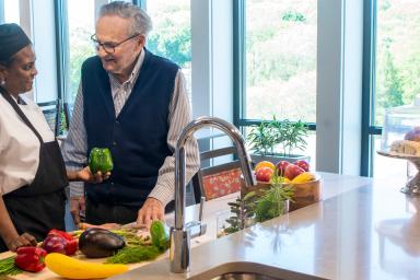 A male resident stands and talks with a female cook while she is holding a green bell pepper.
