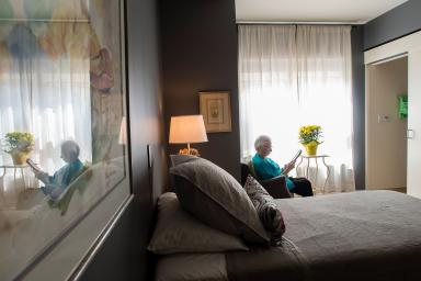 An older adult woman sits in a chair by the window in her room and reads