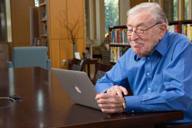 An elder male sits and smiles at a laptop while sitting in a library 