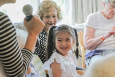 a young girl wearing braids looks up and smiles while a woman speaks into a microphone