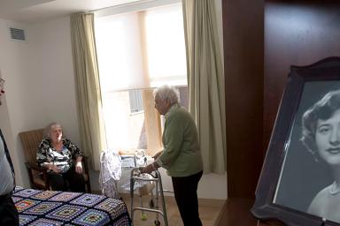 Hebrew SeniorLife's CEO Lou Woolf stands near a residents bed while talking to her and a female friend 