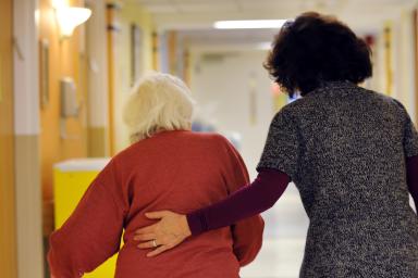 A woman with a walker walks down a hallway with another woman who helps guide her 