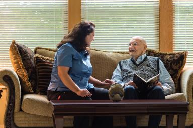 An older male sits on a couch with a woman caregiver and reads.