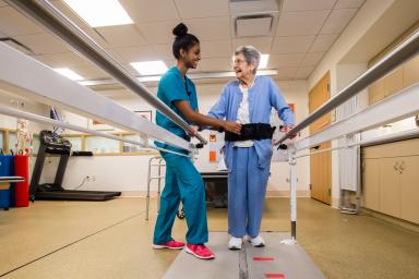 Physical therapist helps older woman walk along balance bars in rehabilitation gym at Hebrew Rehabilitation Center