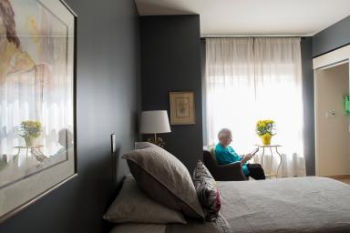 An older adult woman sits in a chair by the window in her room and reads