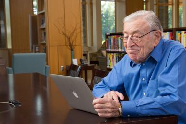An older man smiles while using a laptop.