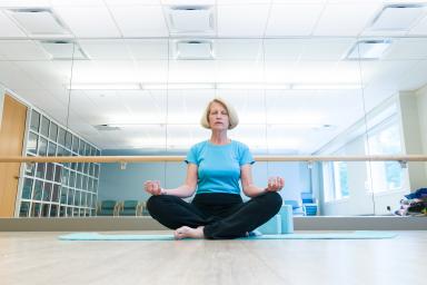 An older woman sits cross-legged and meditating on the floor of a gym at Orchard Cove in Canton, with her eyes closed and hands on her knees.