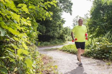 Older man with weights running on gravel path outside with trees.