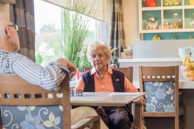 Sitting in a dining area, an older woman in a pink shirt and purple vest talks with a man in a black and white plaid shirt whose back is to the camera.