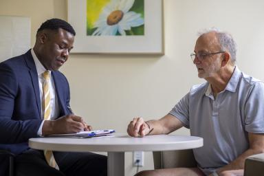 Older man sitting at table with doctor in blue blazer discussing report on clipboard.